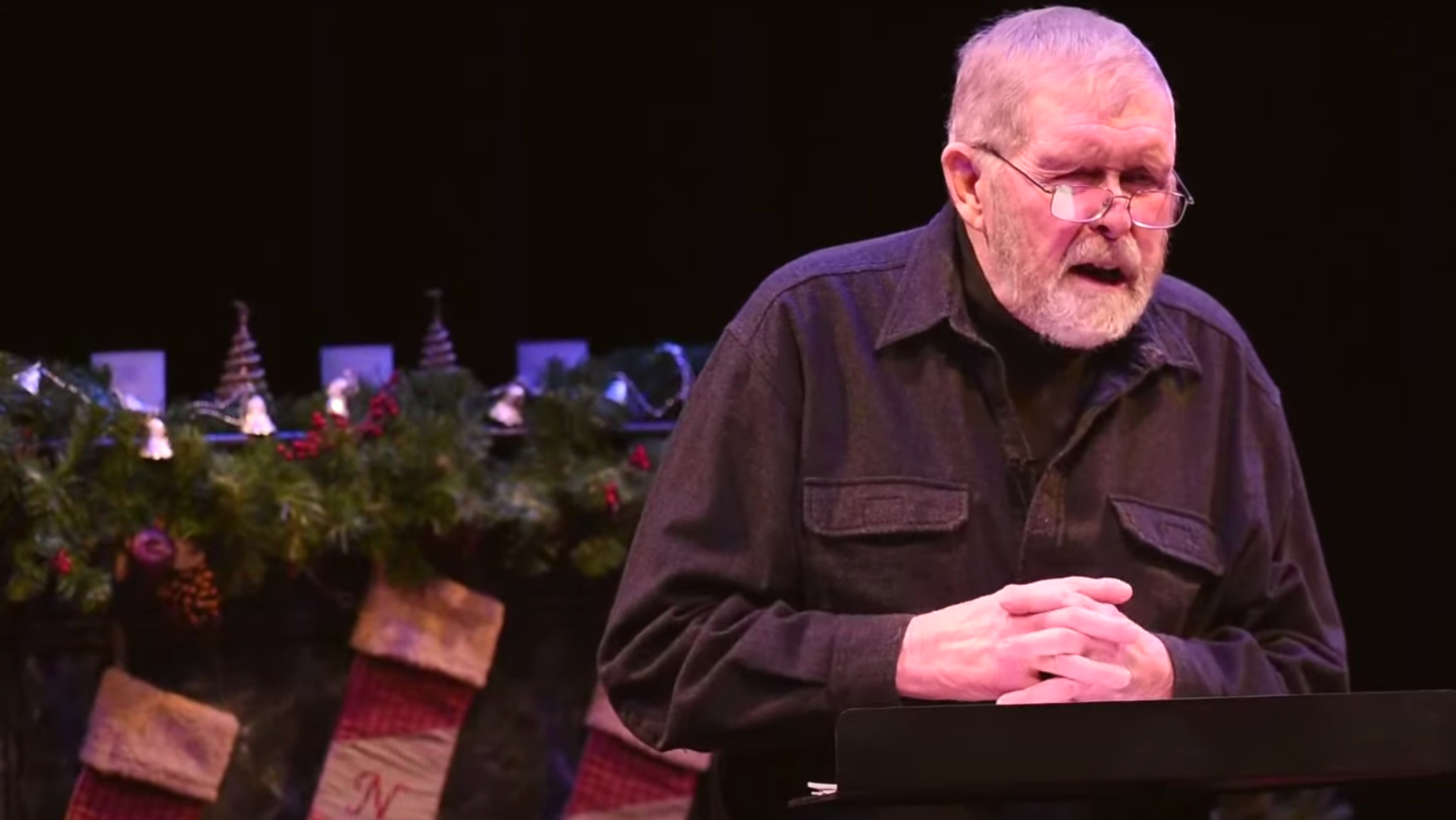 Willem Lange (close up) in performance at LNT in 2020. With his hands folded on his chest, you see him from waist up, and can see a decorated fireplace with stockings (one with the initial L another with initial N, and 3rd with initial T) behind him